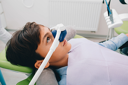 A young person wearing a dental mask and using a dental device, possibly for cleaning teeth or other dental procedures, while seated in a dental chair with a dentist's office setting in the background.