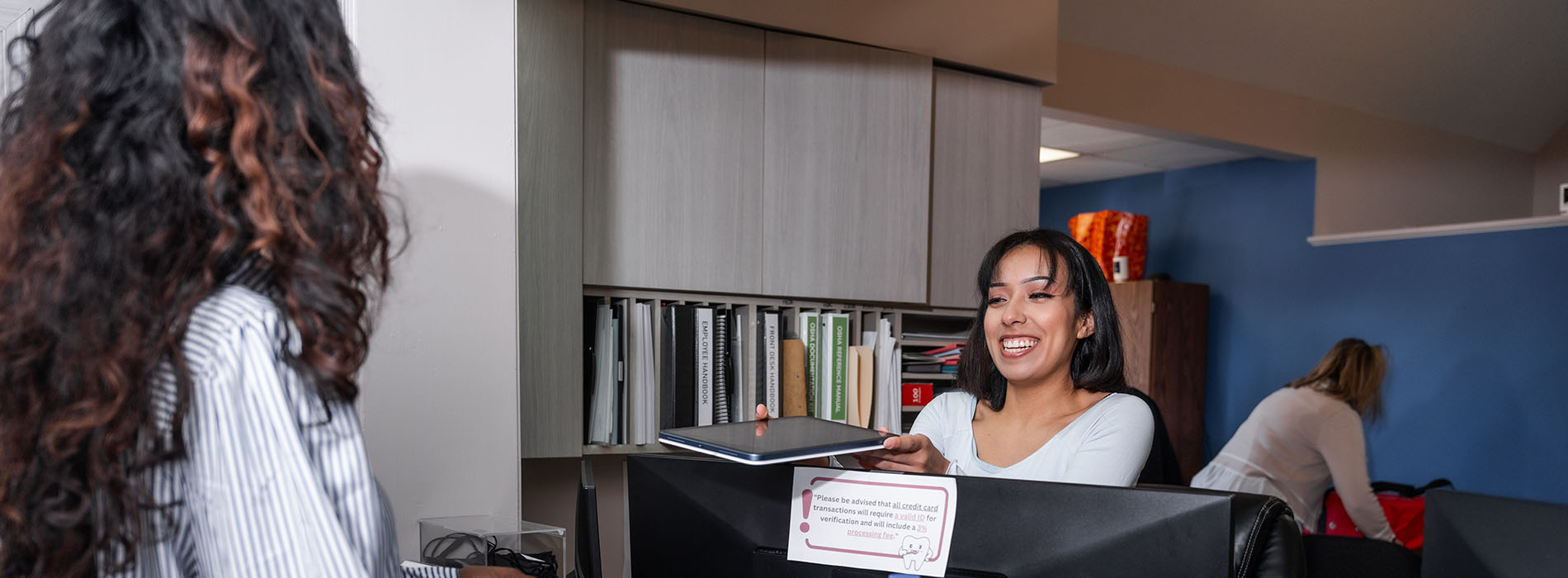 A person standing in front of a desk with a computer monitor, while another person is seated at the desk.