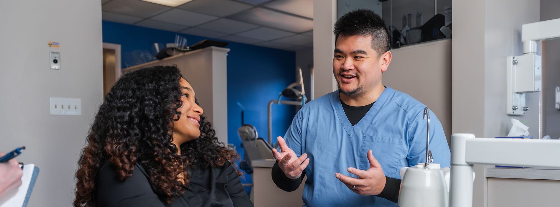 A man and woman are standing next to each other in a room with medical equipment visible in the background, possibly indicating a healthcare setting.
