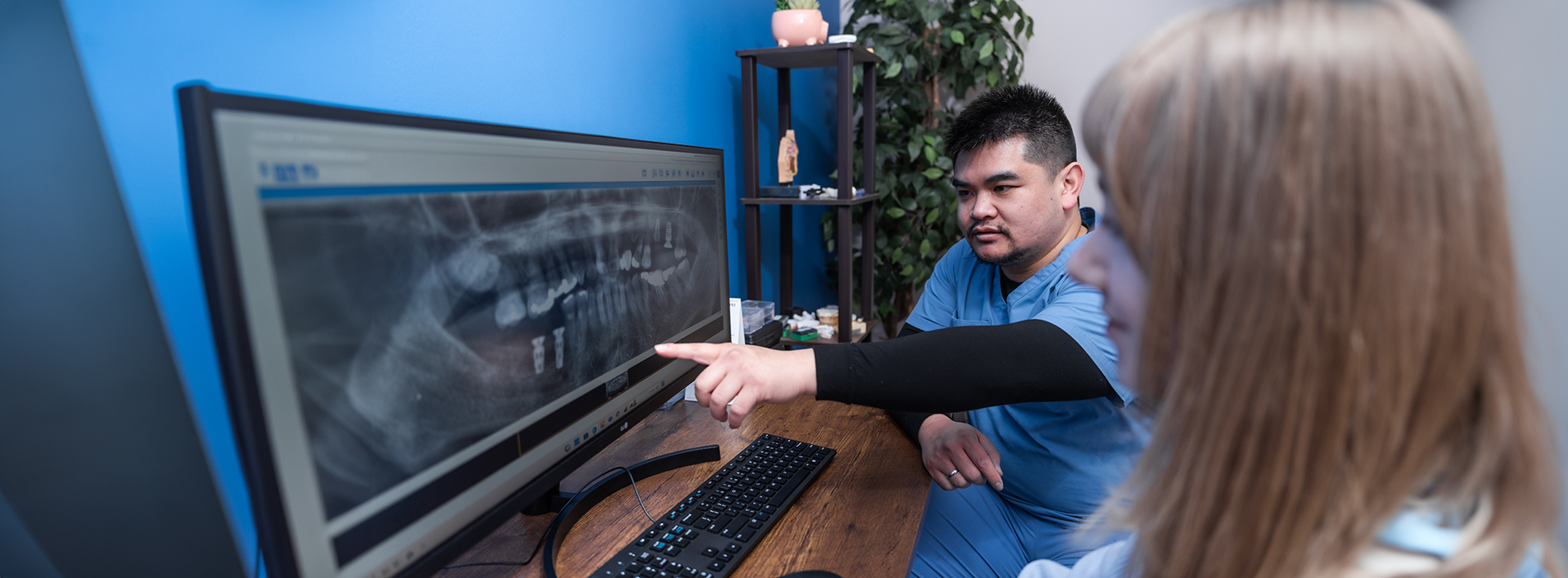 The image shows a person pointing at an X-ray displayed on a computer screen while another individual observes, both are standing in front of a medical imaging device in a clinical setting.