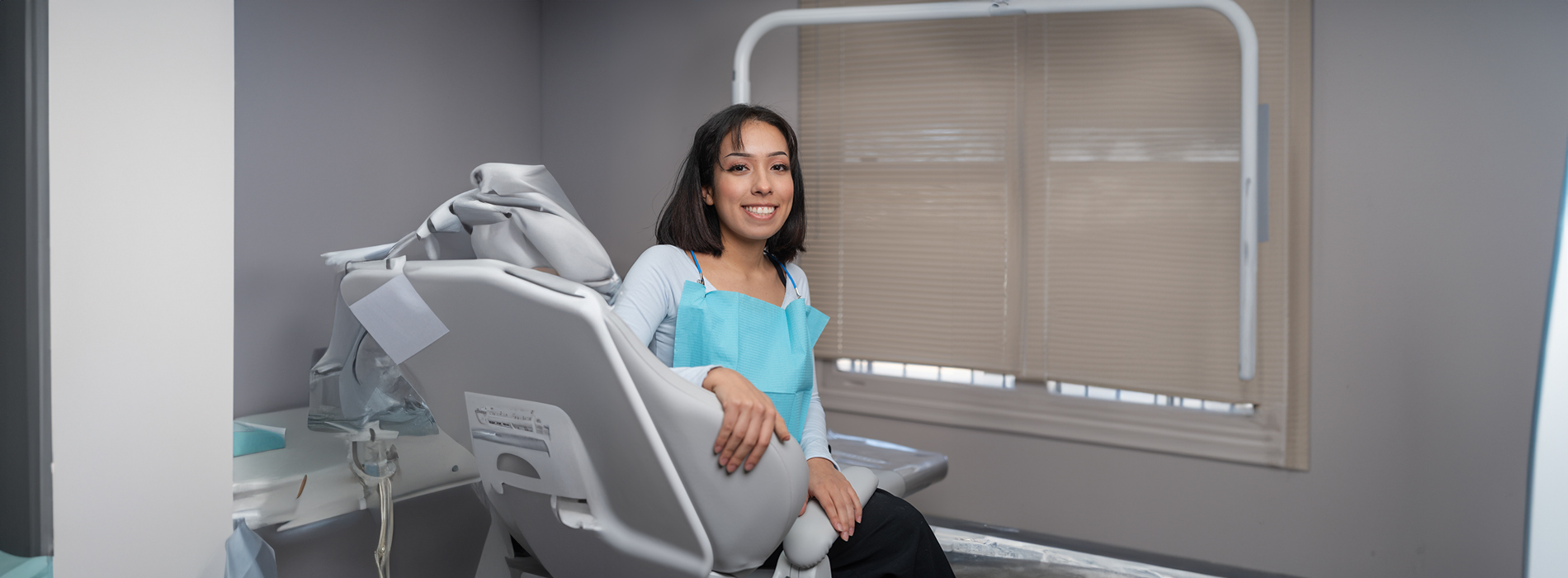 A woman sitting in a dental chair with a smile, wearing a blue shirt, in front of a window with blinds partially drawn.