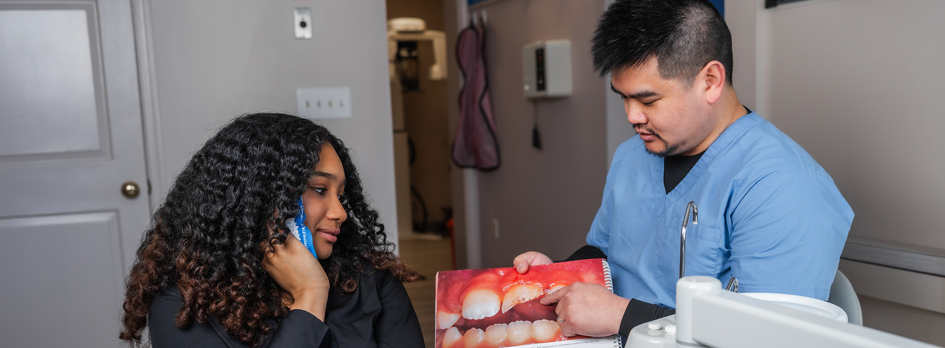A man in scrubs, wearing a mask, is assisting a woman with a medical device while she holds up a box of something red.