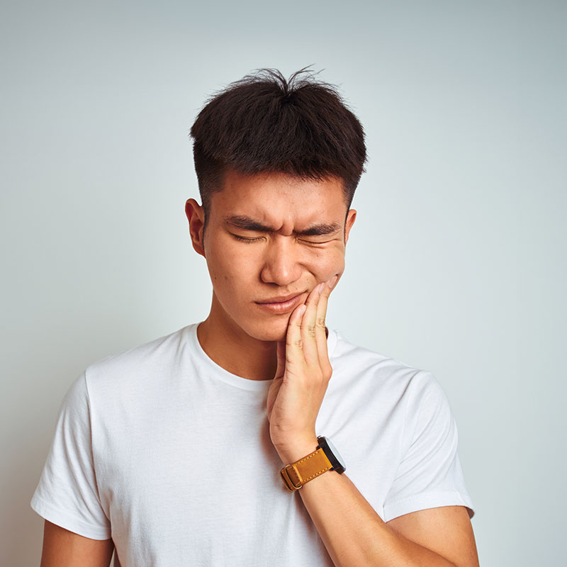 A young man with short hair, wearing a white t-shirt and gold watch, stands against a plain background, making an exaggerated facial expression with his hand on his chin.