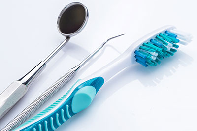 The image shows a collection of dental hygiene tools including a toothbrush with blue bristles, a dental mirror, and dental forceps placed on a white surface with a blurred background.