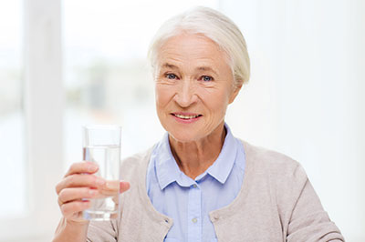 A smiling elderly woman holding a glass of water with both hands, standing indoors.