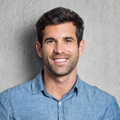 A smiling man with short hair, wearing a light blue shirt, poses against a grey wall.