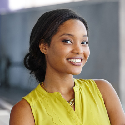 A smiling woman with dark hair wearing a yellow top and posing against a concrete wall.
