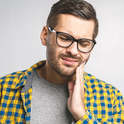 The image shows a man with glasses, wearing a yellow plaid shirt, holding his hand to his chin, expressing concern or contemplation.