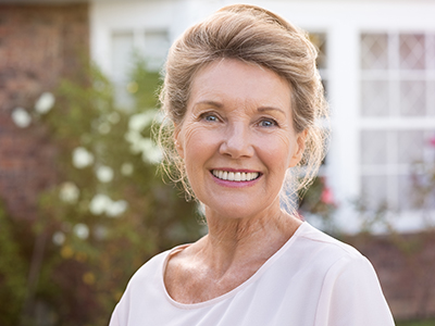 The image shows a smiling older woman with short hair, wearing a pink top, standing outside a house with a brick wall and white flowers.
