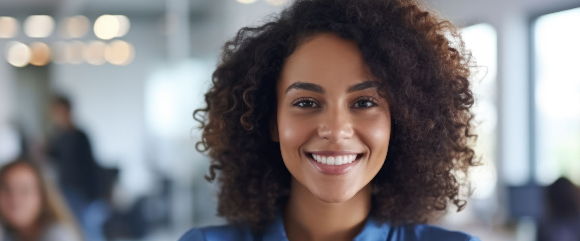 The image depicts a woman with curly hair smiling at the camera, wearing a blue top, set against an indoor office environment.