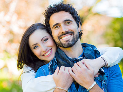A man and woman are hugging each other, smiling, with the man wearing a scarf around his neck.
