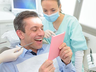 The image shows a man smiling at a pink card he's holding while sitting in a dental chair with a woman dentist attending to him, both wearing surgical masks and gloves.